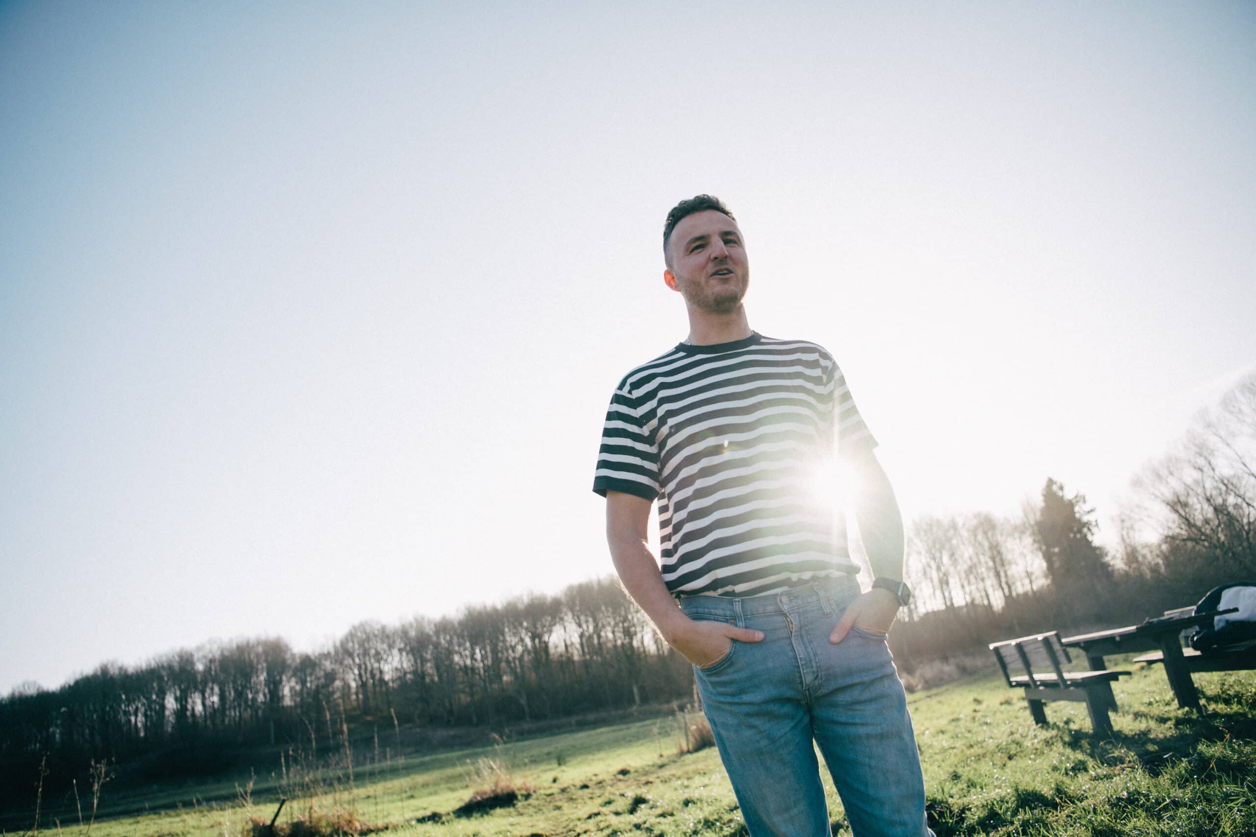 This image shows Edis standing outdoors in a field, wearing a striped t-shirt and jeans. The sun shines brightly behind him, creating a glowing halo effect and lens flare. In the background, there are trees, a picnic table, and an open grassy area under a clear sky. The scene feels peaceful and natural.