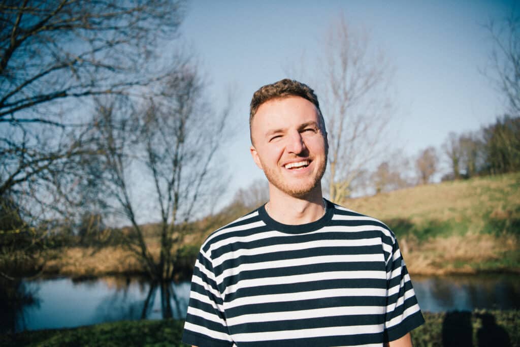 This image shows Edis standing outdoors, smiling warmly in the sunlight. He is wearing a striped shirt, and behind him is a serene natural setting with trees, a calm body of water, and grassy hills under a clear blue sky. The atmosphere is peaceful and cheerful.