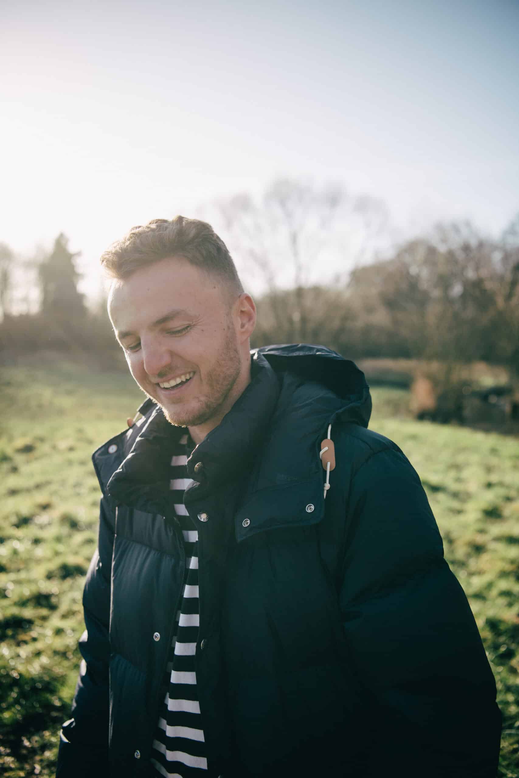 Edis 13 This image shows Edis outdoors, smiling warmly and looking slightly down. He is wearing a striped shirt under a dark puffer jacket, and the sunlight behind him creates a soft glow. The background features a grassy field and trees, giving the scene a peaceful and relaxed atmosphere.