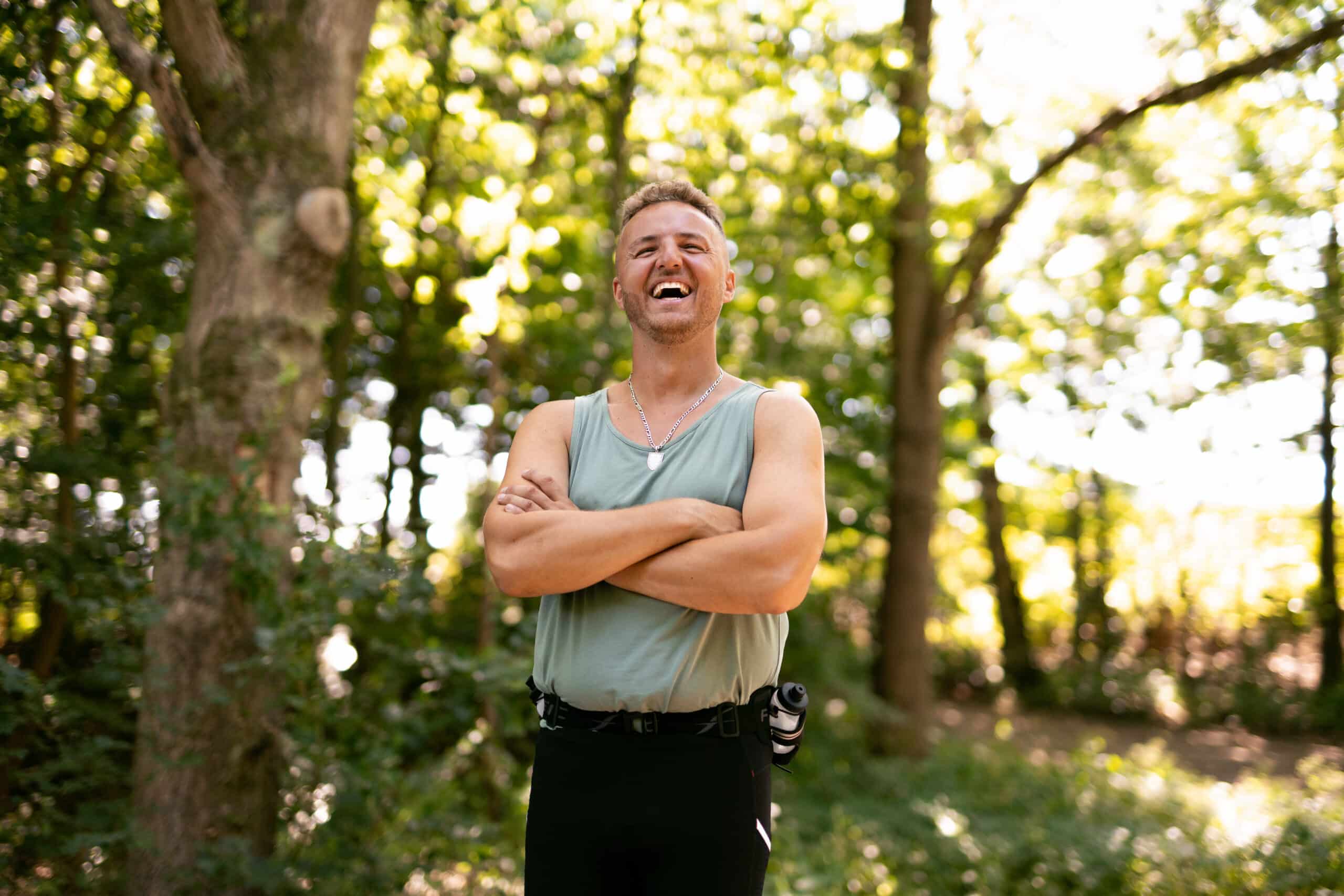 Edis_griner_med_krydsede_arme_i_skov_med_gra_top_bredformat_50 Edis smiling and standing in a forest, wearing a light green tank top and black pants with a hydration bottle attached to his waist. He has his arms crossed and is illuminated by soft sunlight filtering through the trees.