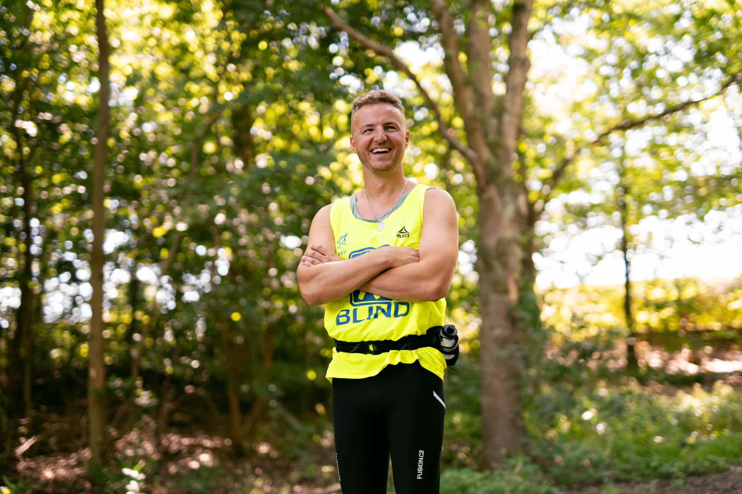 Edis_griner_med_krydsende_arme_i_skov_med_gul_vest_bredformat_50 Edis running along a sunlit path surrounded by greenery, wearing a bright yellow vest with the word 'BLIND' and an icon symbolizing blindness. He is smiling, focused, and appears confident as he enjoys the run.
