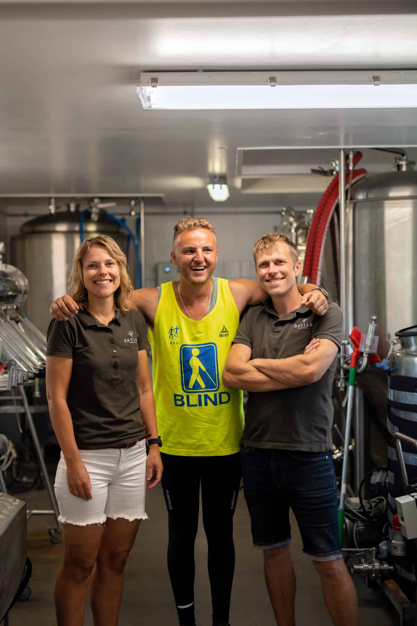 Edis is standing in the center of a brewing facility, smiling brightly with his arms around two people. He is wearing a bright yellow vest with the word Blind and black athletic leggings. To his left is a woman in a dark polo shirt with white shorts, smiling, and to his right is a man in a dark polo shirt and jeans, also smiling with his arms crossed. Stainless steel brewing equipment and red hoses are visible in the background.