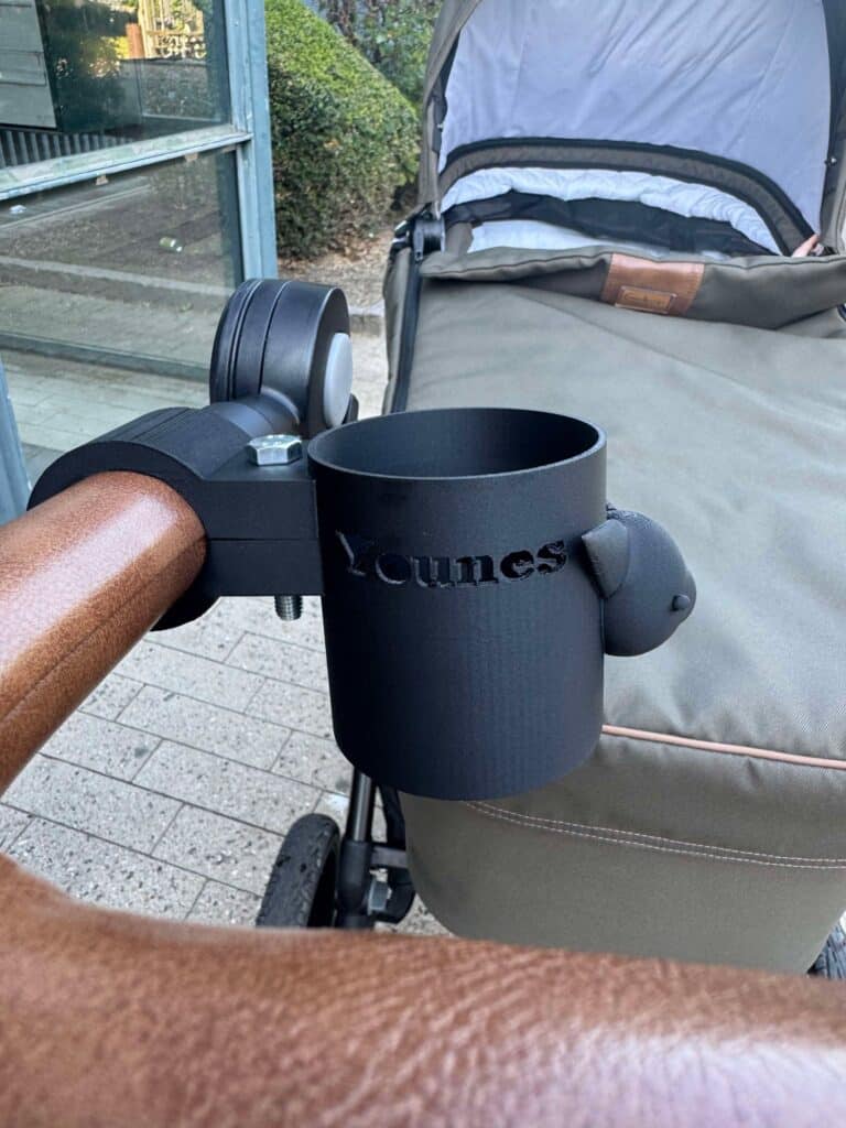 This image shows a stroller handle with a custom 3D-printed cup holder attached. The cup holder is black and has the name "Younes" embossed on its side. The stroller has a brown and beige color scheme, with a visible seat or bassinet in the background. The attachment point appears to be secured with a bolt, and the cup holder is mounted to a rounded handlebar, likely for convenience during walks.