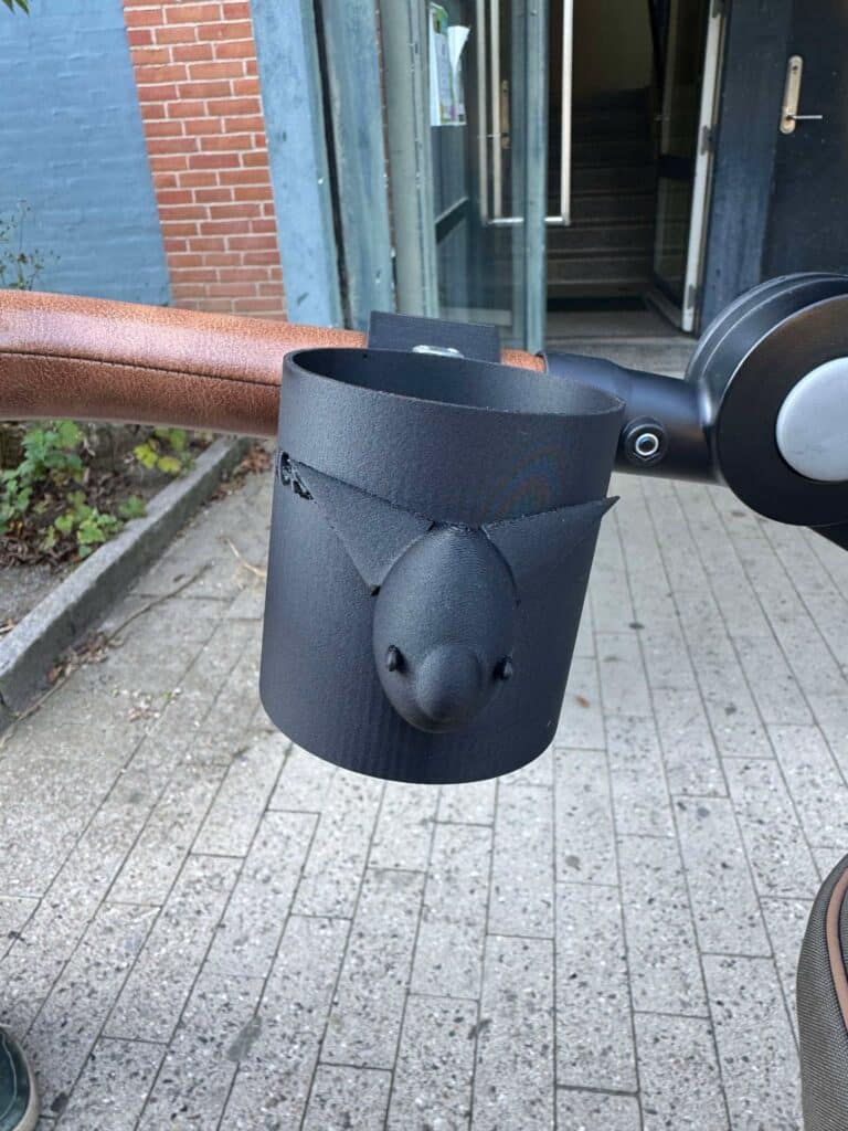 This image shows a close-up view of a black, 3D-printed cup holder attached to a stroller handle. The cup holder features a decorative fox head design, with pointed ears and a snout in the center. The holder is secured to a brown handlebar, and the attachment mechanism involves a bolt. The background includes a tiled pathway and an entrance to a building with a partially open door.