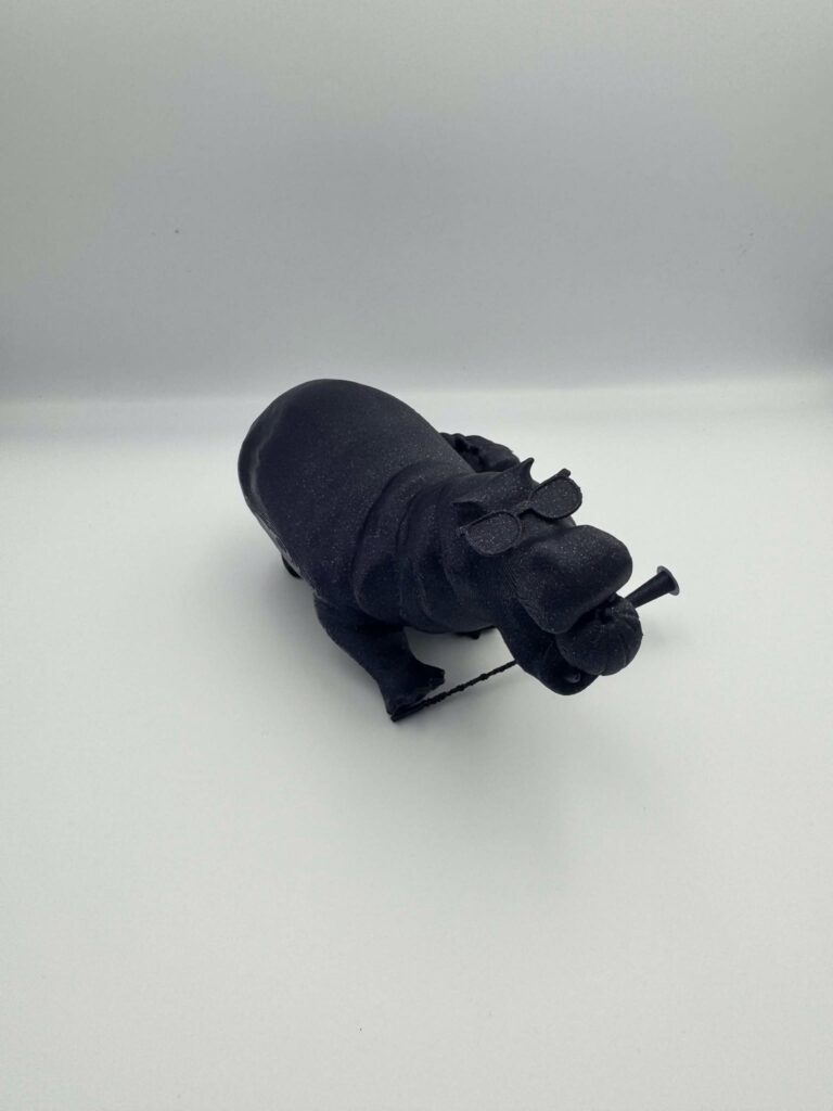 This image depicts a 3D-printed model of a black hippopotamus with a playful and creative design. The hippopotamus is wearing small glasses, adding a humorous touch, and is holding an object resembling a string or rope in its mouth. The figure has a matte texture and rounded body details, capturing the essence of a hippo while adding whimsical elements to its appearance. The overall design is imaginative and charming.