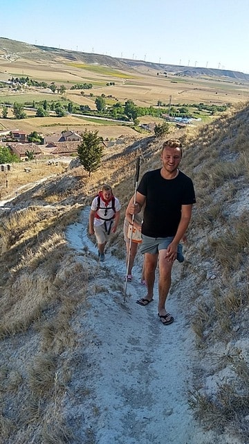 Hiking Through Scenic Hills with Open Fields in the Background The image shows two hikers walking along a narrow dirt trail on a hillside. The person in the foreground is Edis. He is smiling and holding a white cane, wearing a black t-shirt and shorts. Behind him, another hiker with a backpack is following. The hillside is dry with sparse vegetation, and in the background, a scenic view of green fields, scattered buildings, and wind turbines on the horizon is visible. The setting evokes a sense of adventure and connection with nature.