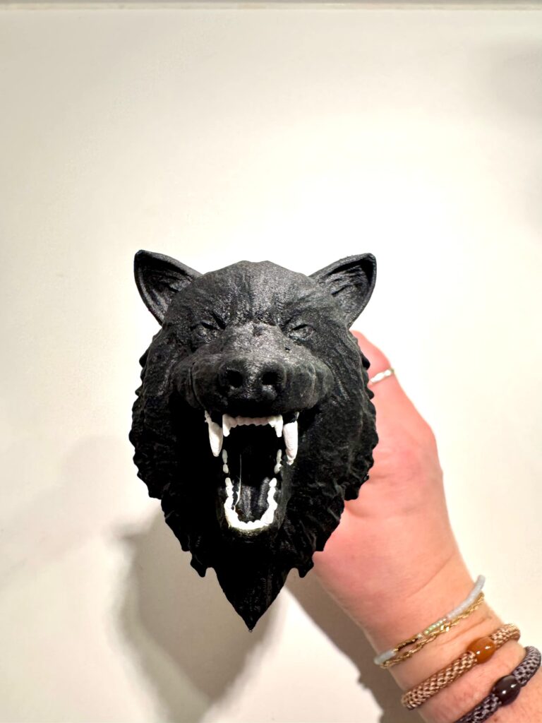A close-up view of a black wolf-shaped knife pommel, featuring detailed fur texture and an open mouth with prominent white fangs. The handle is held by a hand wearing several bracelets on a white background.