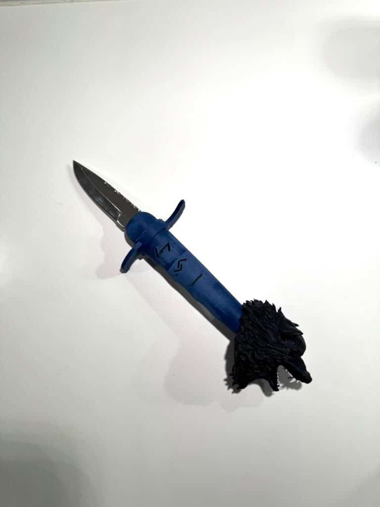 A knife with a blue handle featuring engraved grey runes and a black wolf-shaped pommel, placed on a clean white surface. The blade is partially extended, showcasing its metallic finish.