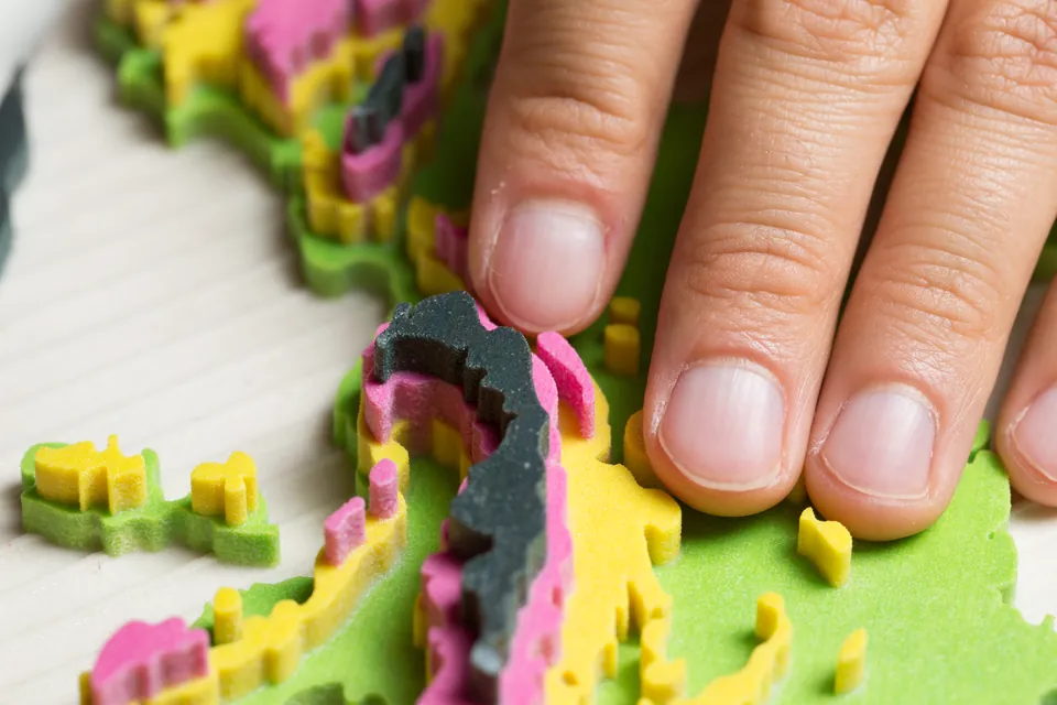 Close-up of fingers exploring a raised relief map. The land is built up in layers of green, yellow and pink, with a dark grey coastline standing out under the fingertips.