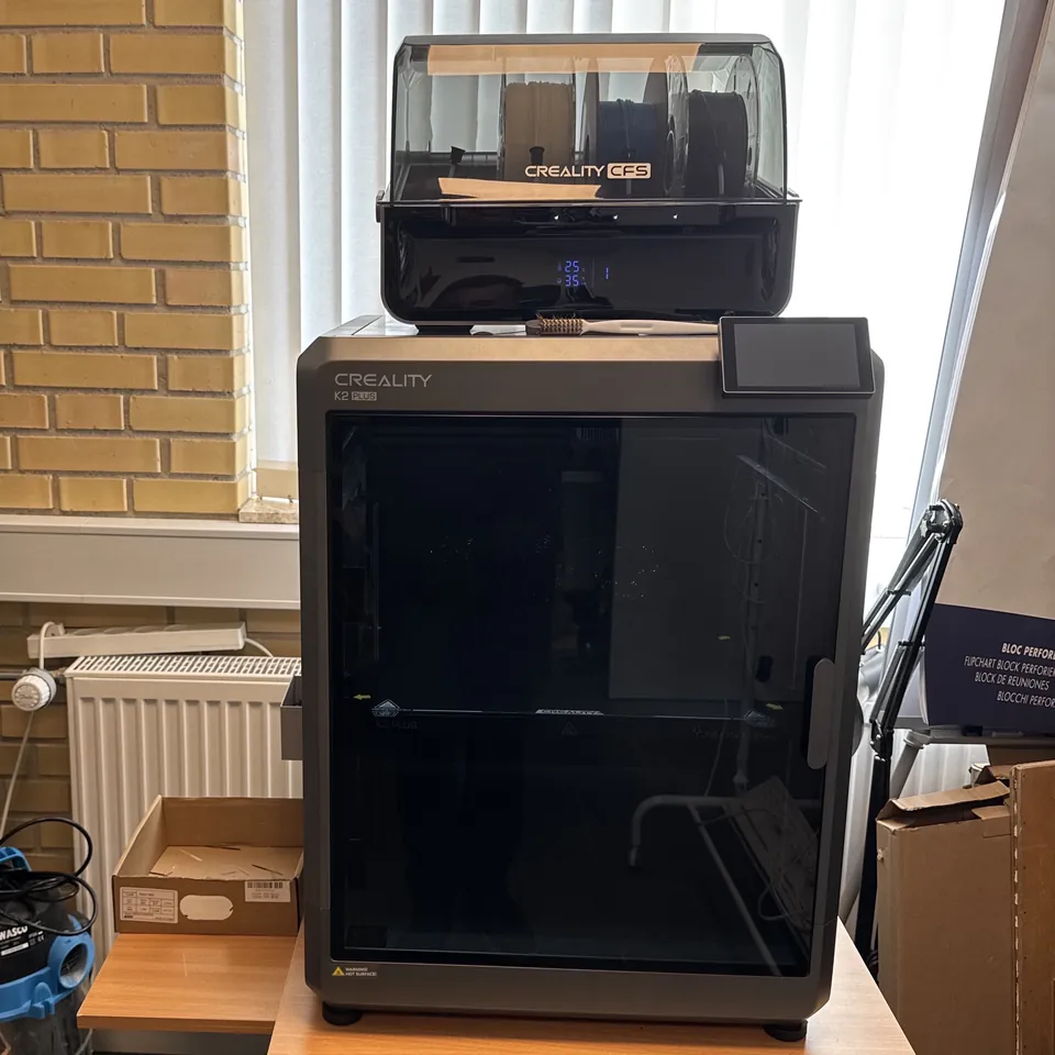 A Creality K2 Plus 3D printer on a wooden table in a workshop, in front of a window and a yellow brick wall: a tall, dark grey, fully enclosed cabinet with a glass door and a small touch screen, with the CFS filament box on top holding three spools.