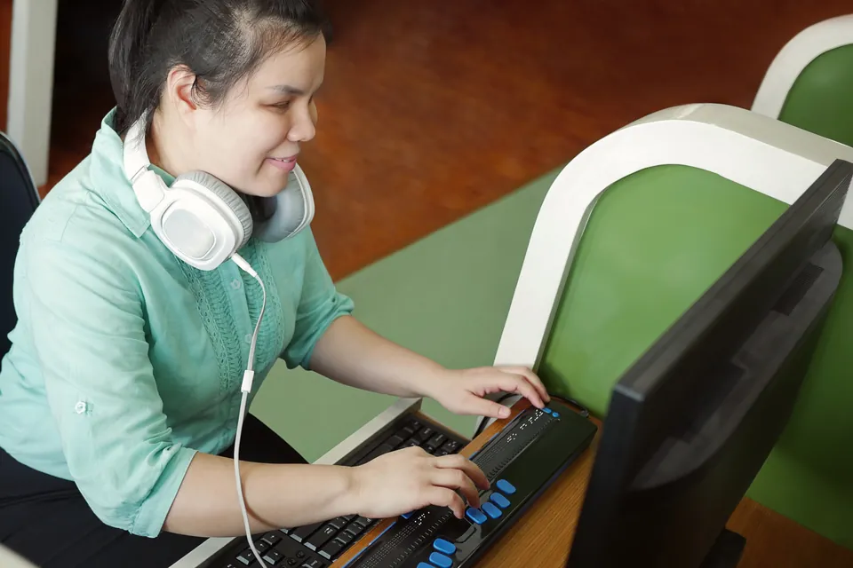 A young blind woman with white headphones around her neck works at a computer, her fingers resting on a refreshable braille display in front of the keyboard.
