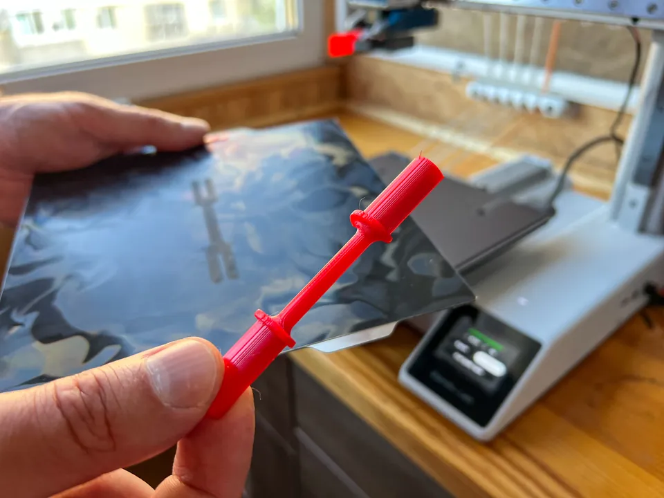 A hand holding up a small red 3D printed part, a thin rod with two raised rings and a thicker handle at the end. Behind it, a black flexible build plate and a white printer on a wooden table.