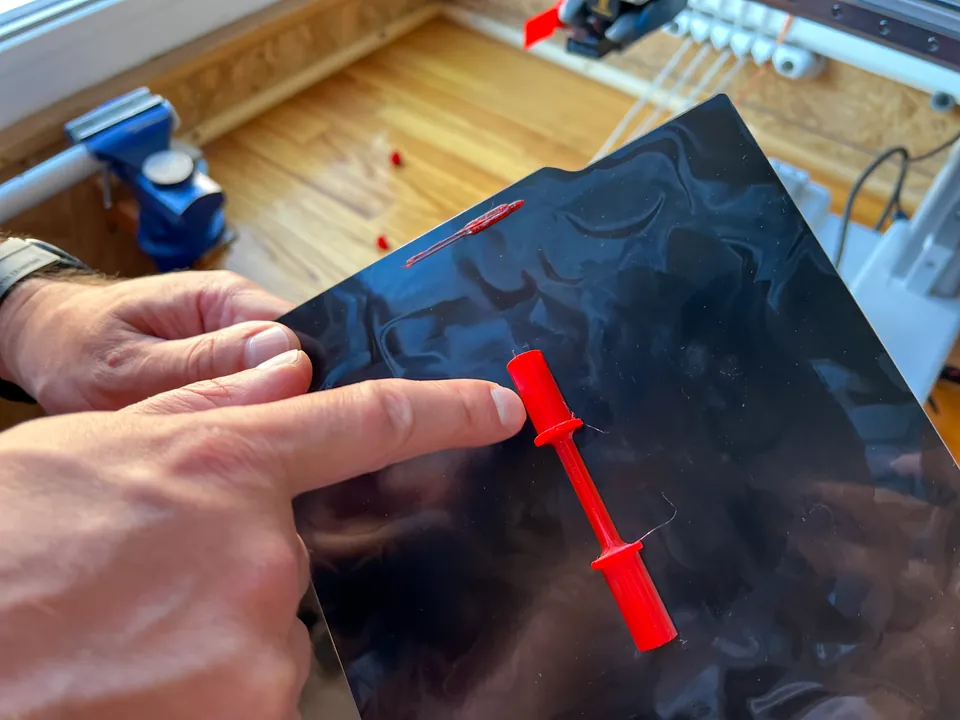Fingers touching small red printed parts that are still lying on a black flexible build plate, with the printer in the background.