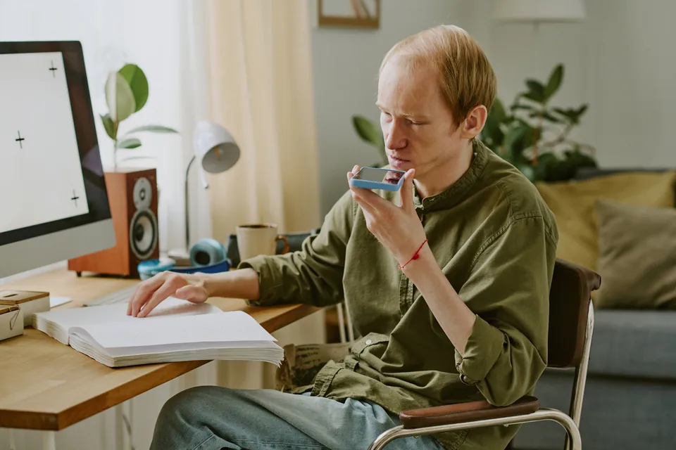 A blind man sits at a desk at home and speaks into his phone, with one hand resting on an open braille book.