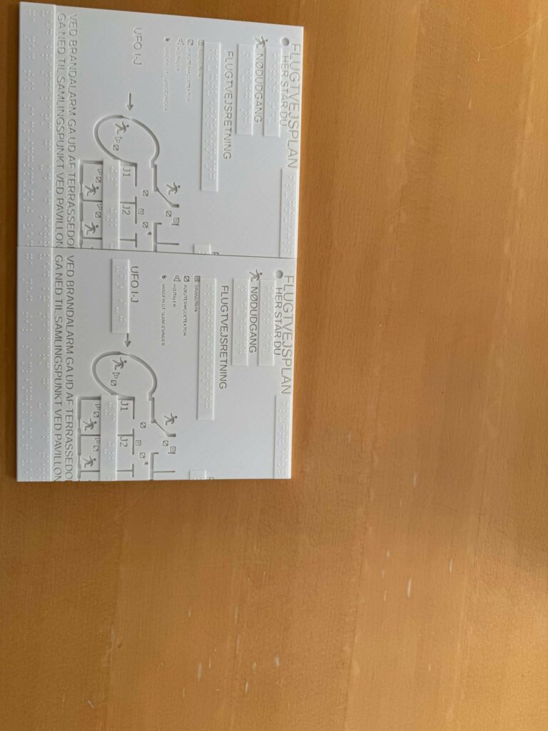 A tactile evacuation plan is printed on a white surface with engraved routes, symbols, text and Braille labels for emergency exits and assembly points.