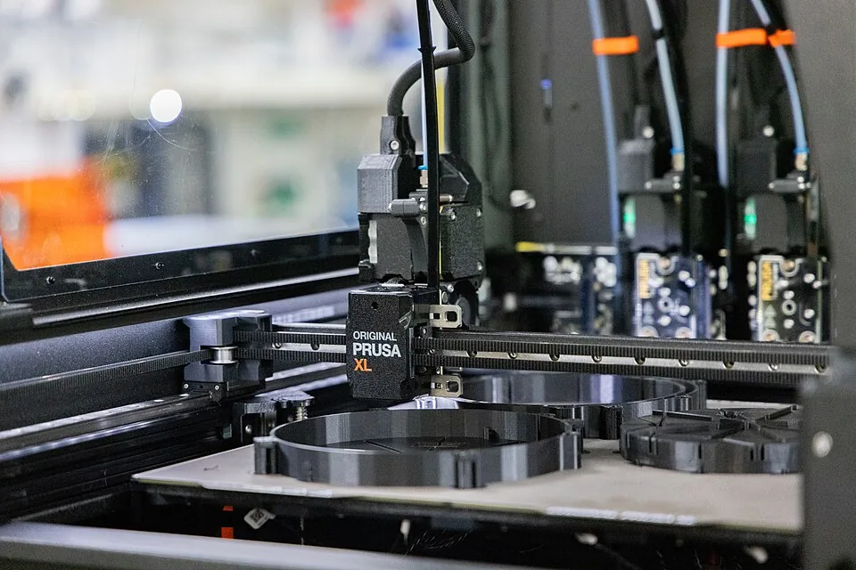Et printhoved på Prusa XL, der printer store runde sorte dele på pladen, mens to andre printhoveder venter i docken bagved med deres blå og hvide filamentrør mærket med orange clips.