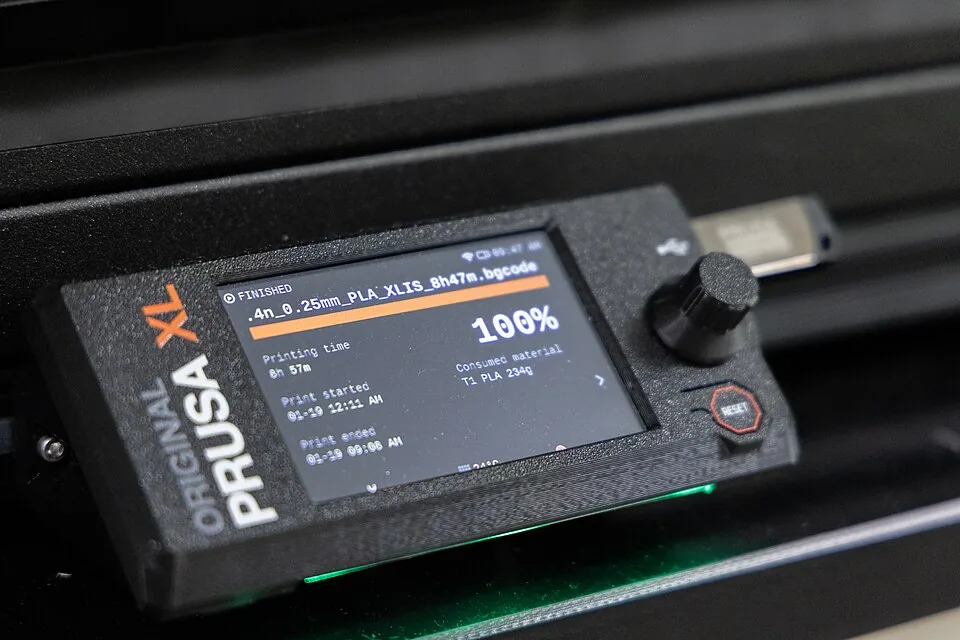 Prusa XL's betjeningsskærm, der viser et færdigt print på 100 procent efter 8 timer og 57 minutter, med en rund drejeknap ved siden af skærmen.