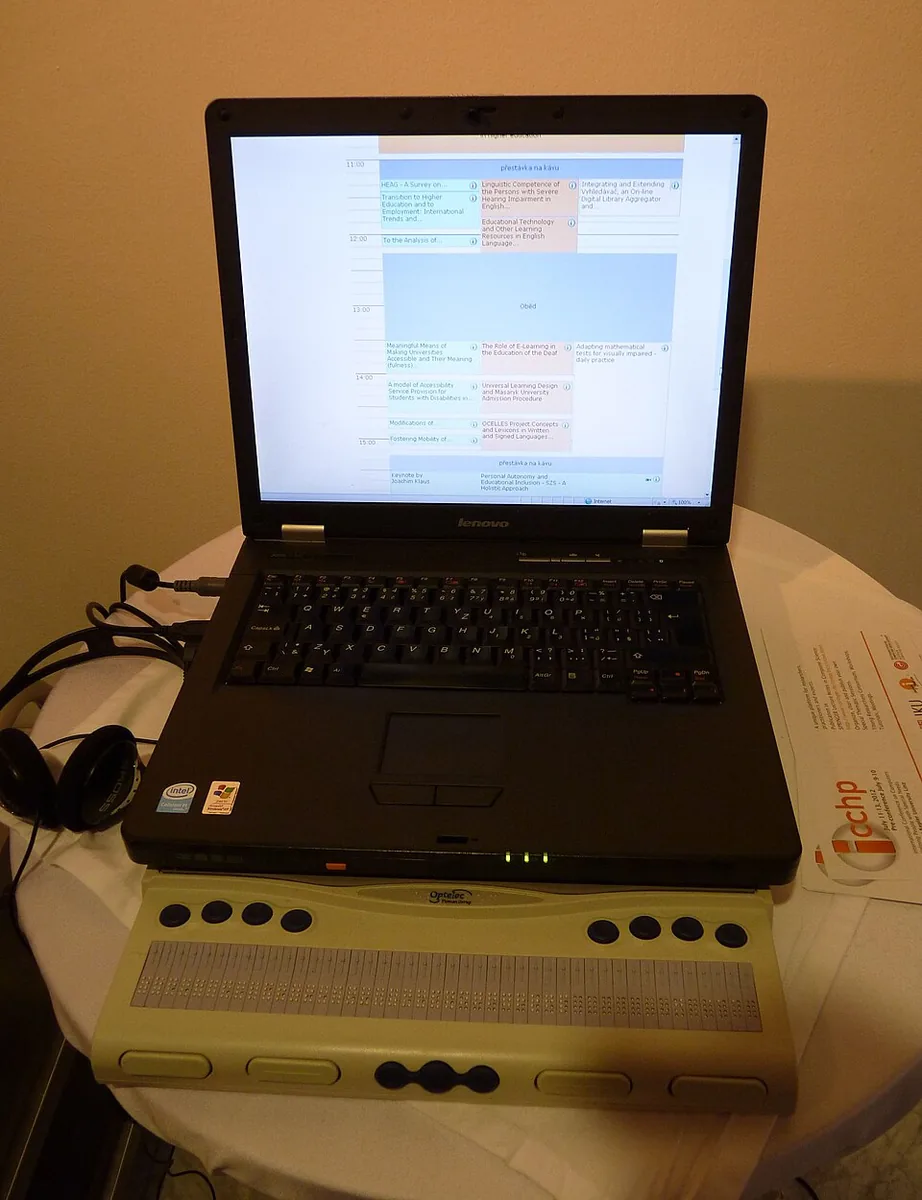 En bærbar computer, der viser en webside, med et blåt punktskriftdisplay foran tastaturet og et par hovedtelefoner ved siden af.