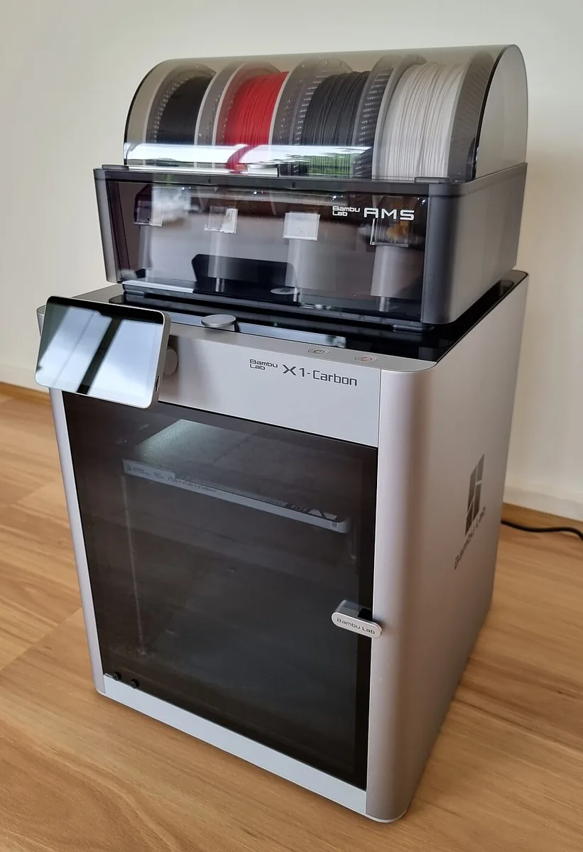 En Bambu Lab X1 Carbon 3D-printer på et trægulv: et sølv- og sort kabinet med en tonet glaslåge og en lille berøringsskærm, med AMS-filamentenheden ovenpå med fire spoler, sort, rød, mørkegrå og hvid.