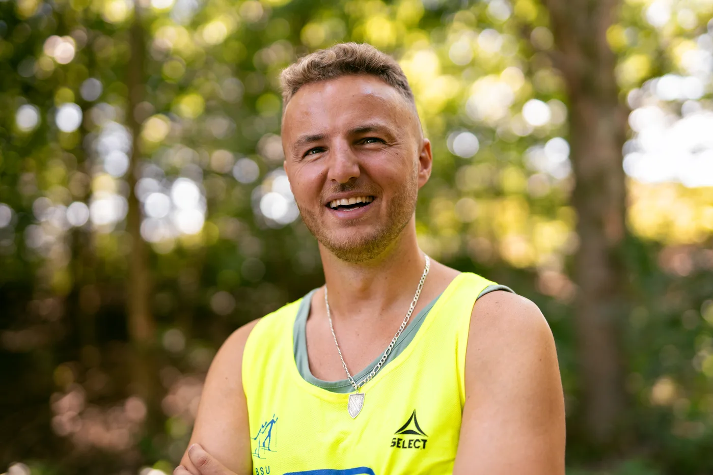 A friendly head-and-shoulders portrait of Edis smiling in woodland, wearing a bright yellow high-visibility vest and a silver chain, with green trees blurred behind him.