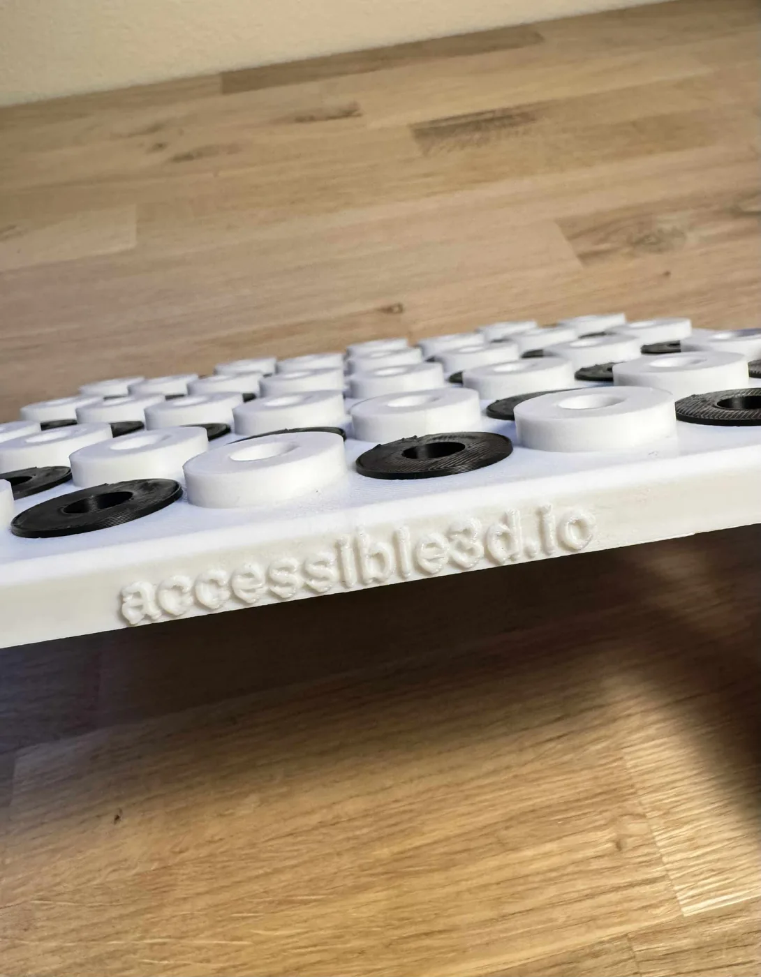 A white 3D-printed tactile chess board sits on a wooden surface, with black and white disc-shaped playing pieces seated in raised circular holes and accessible3d.io embossed along the front edge.