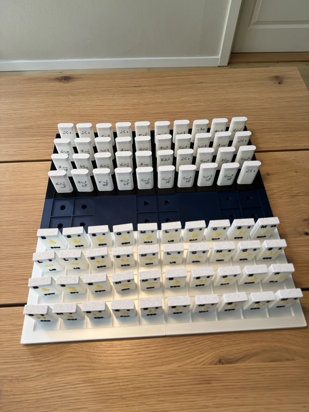 A 3D-printed tactile Stratego board sits on a wooden surface, with a navy and white grid and two armies of upright playing pieces marked with Braille dots and short raised letters.