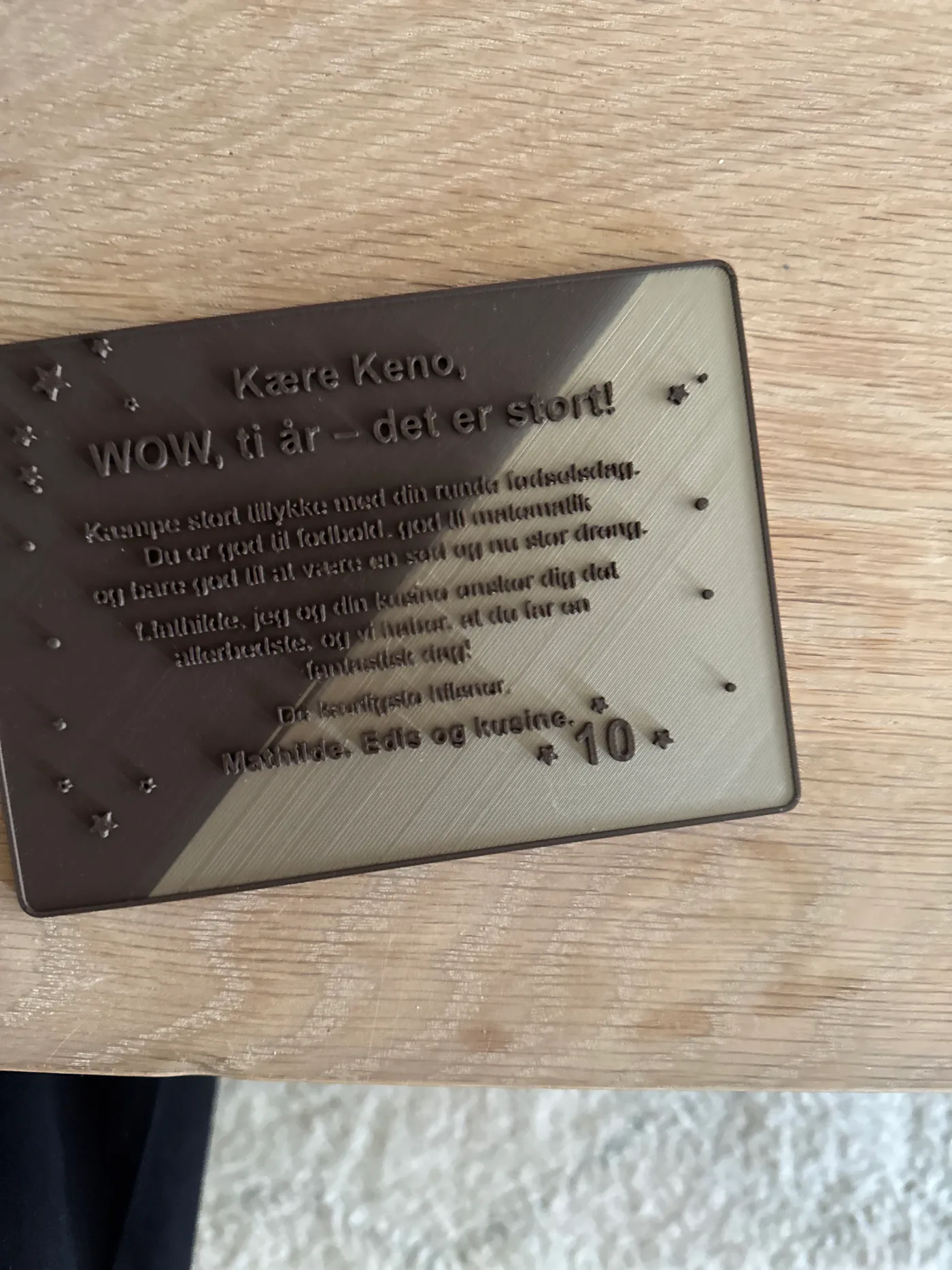 En 3D-printet fødselsdagskortplade med en tofarvet diagonal deling og hævet dansk tekst, printet uden support.