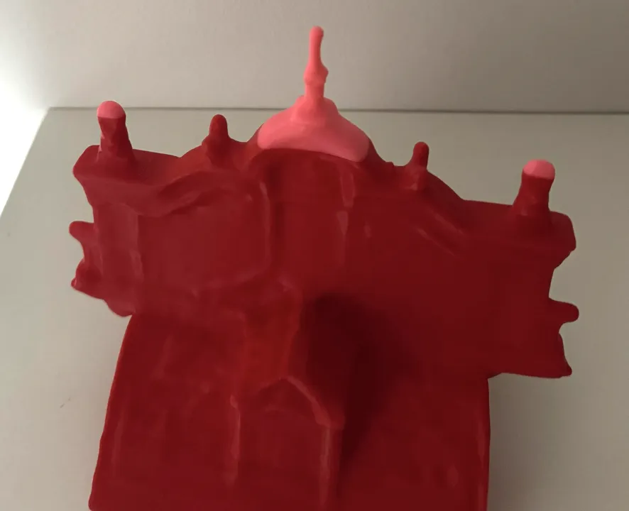 Photo of the 3D printed Børsen model in red with a pink central spire and pink tower tips, standing on a light shelf against a grey wall.