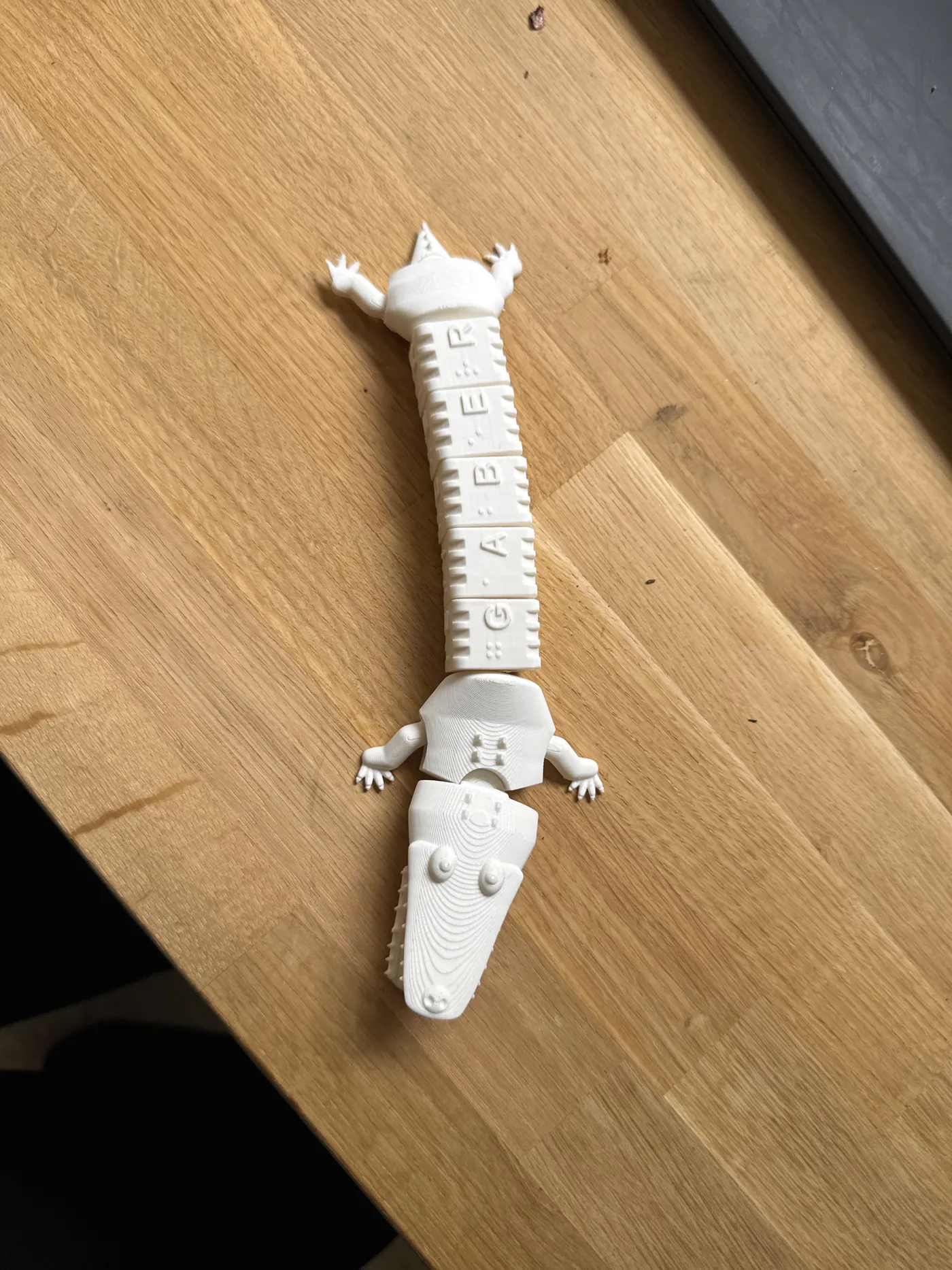 Top-down photo of the white 3D printed word crocodile on an oak table, its five turnable body segments spelling gaber in braille dots and raised capital letters, with clawed legs and a finned tail.