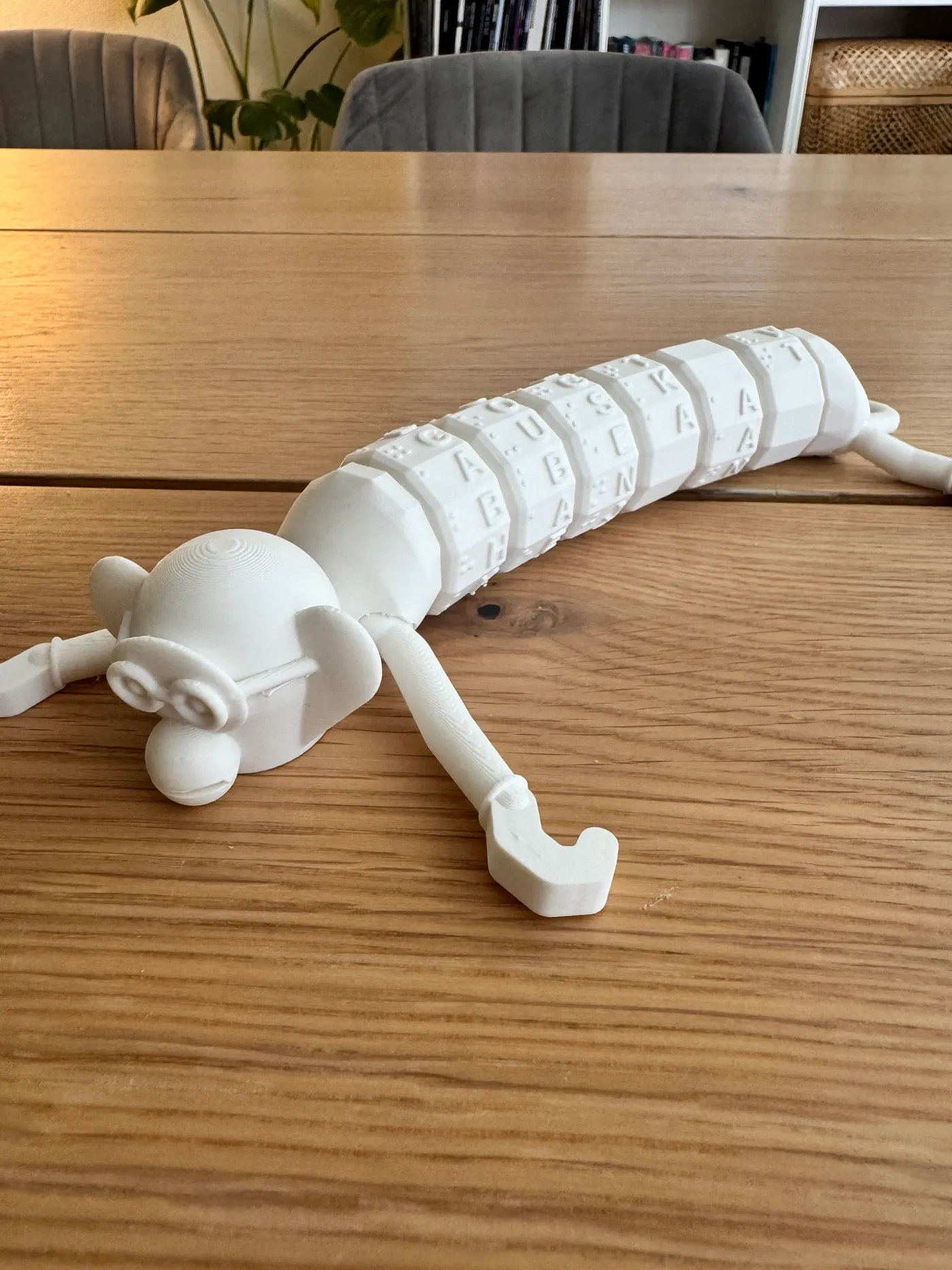 Den hvide 3D-printede brailleabe hænger på kanten af en skabslåge i træ i én krogehånd, hovedet med runde taktile briller vendt udad og bogstavperlerne op langs kroppen.