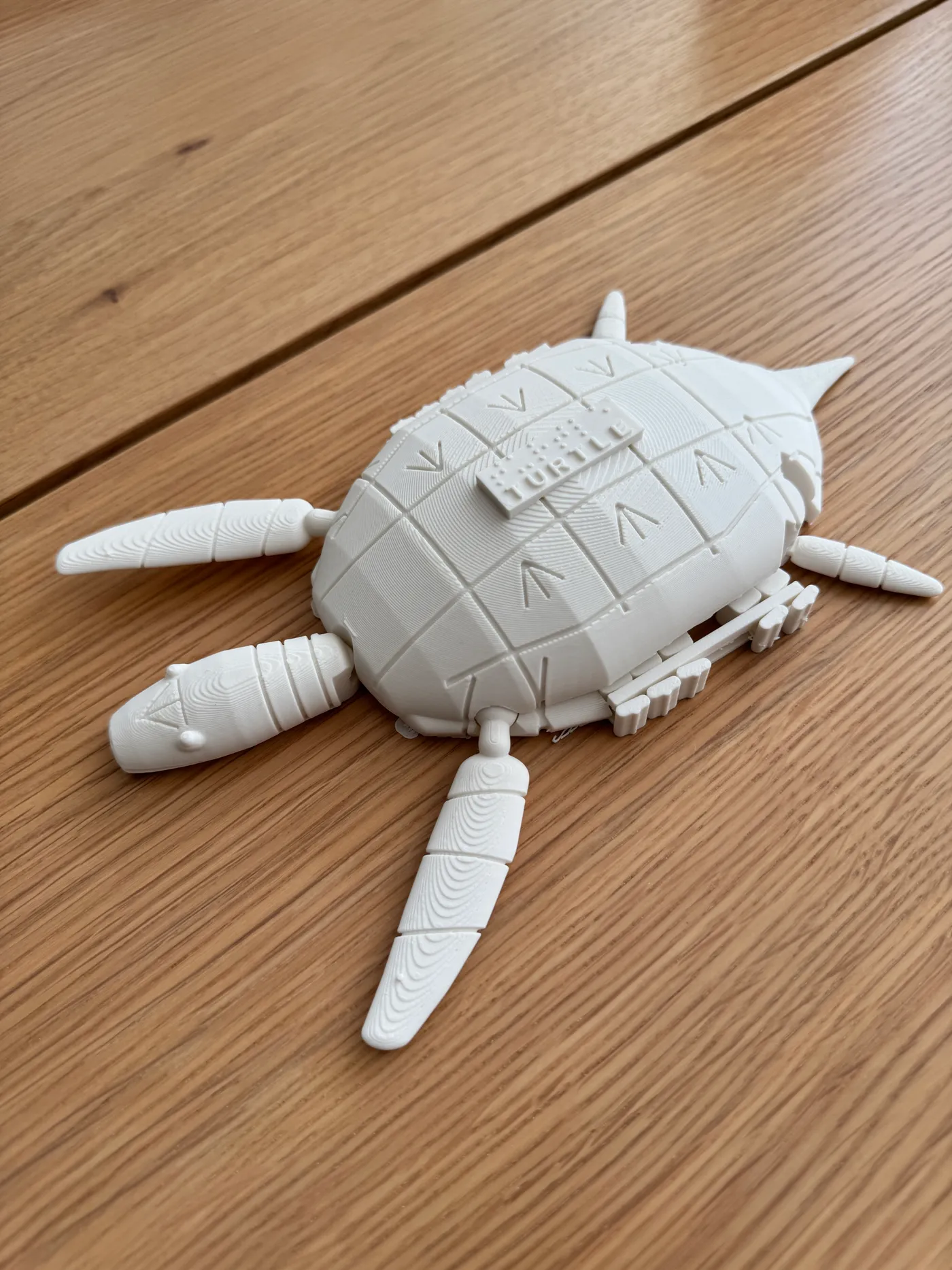 Frontfoto i perspektiv af den hvide 3D-printede braille-havskildpadde med tælleramme på et træbord, med bevægelige luffer, glidebrikker i skjoldkanten og ordet TURTLE i punktskrift.