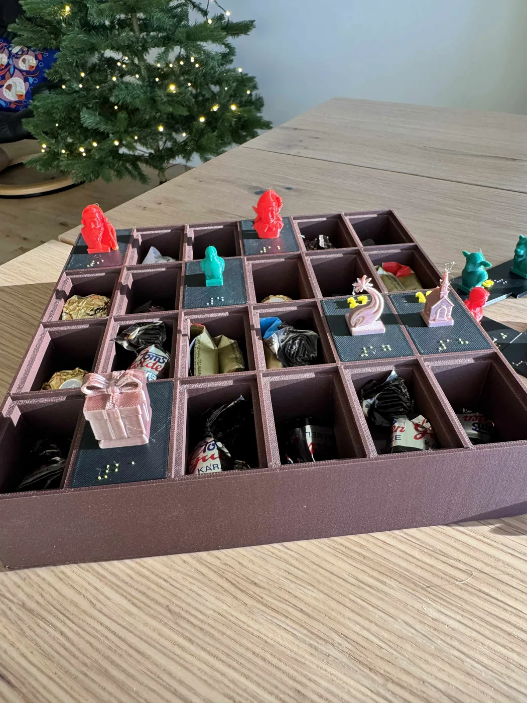 Set ovenfra af en 3D-printet punktskrift-julekalender på et træbord, med små rum, hævede punktskrifttal, farverige pyntebuster og foliepakket chokolade.