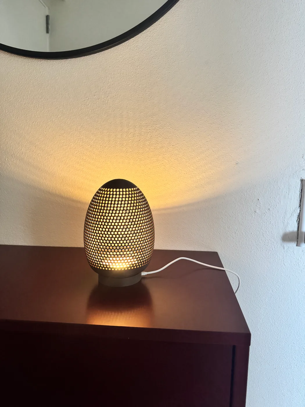 A 3D-printed egg lamp sits on a wooden table, with a warm glow shining through its perforated shell.