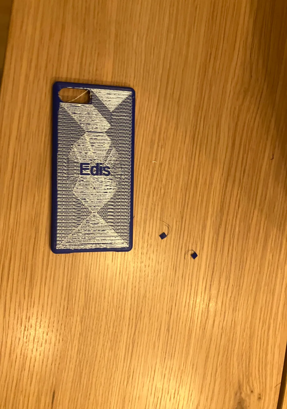 Photo of the blue 3D printed iPhone SE case lying on a wooden table, its back showing a white ironed diamond pattern with the name Edis in raised blue letters.