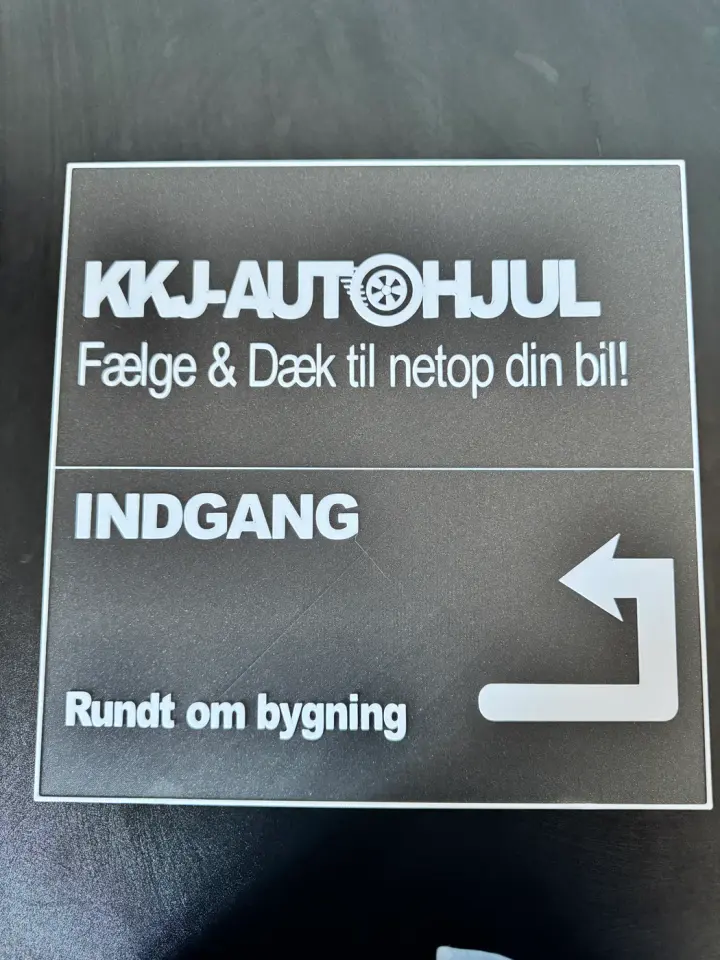 A large 3D-printed workshop directional sign shows high-contrast white raised text on a dark textured background, displaying the brand KKJ-AUTOHJUL.
