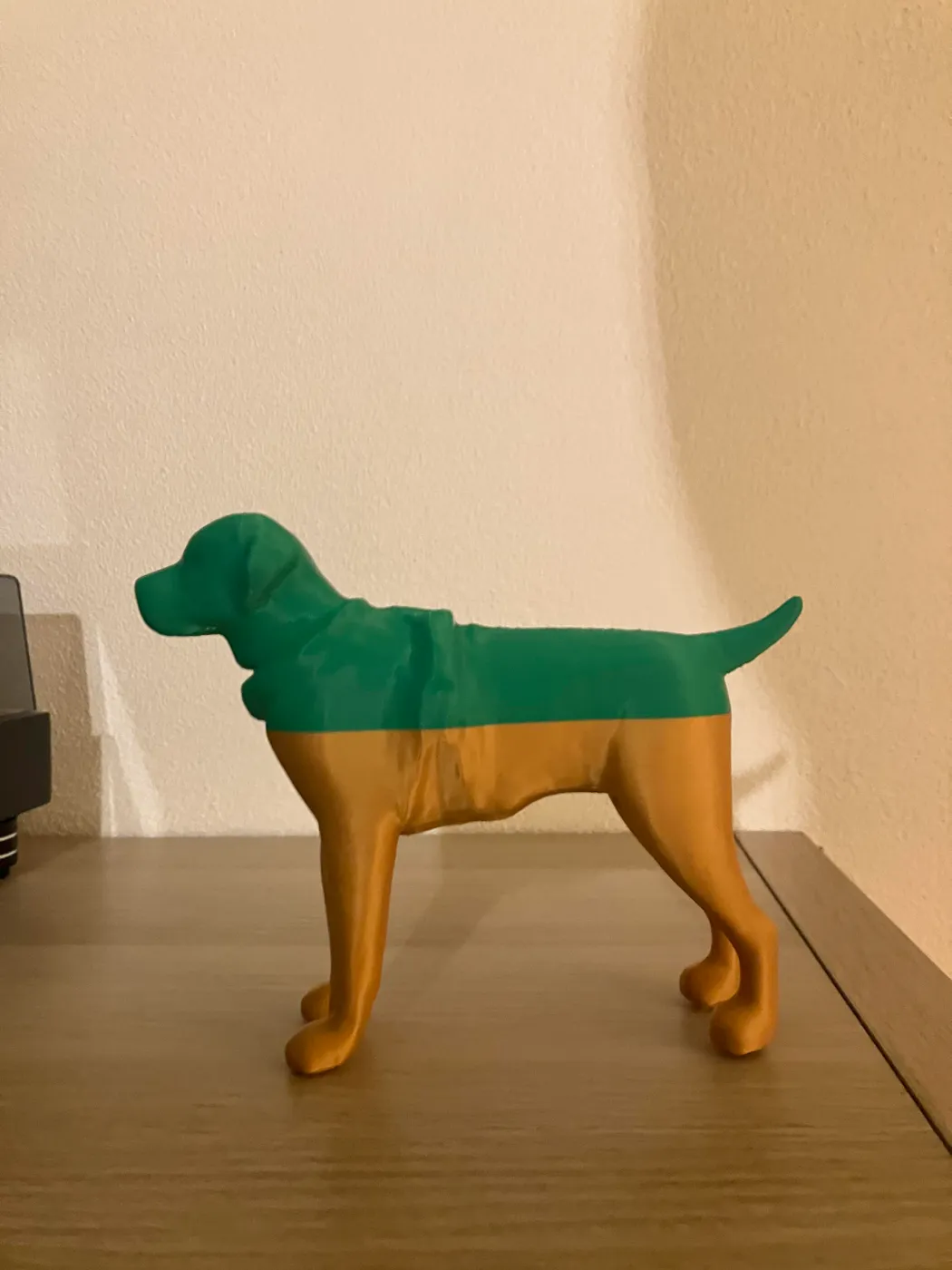 Photo of the 3D printed Labrador guide dog figure standing on a desk in profile, its head, chest and back printed in green and its legs and lower body in orange, tail relaxed.