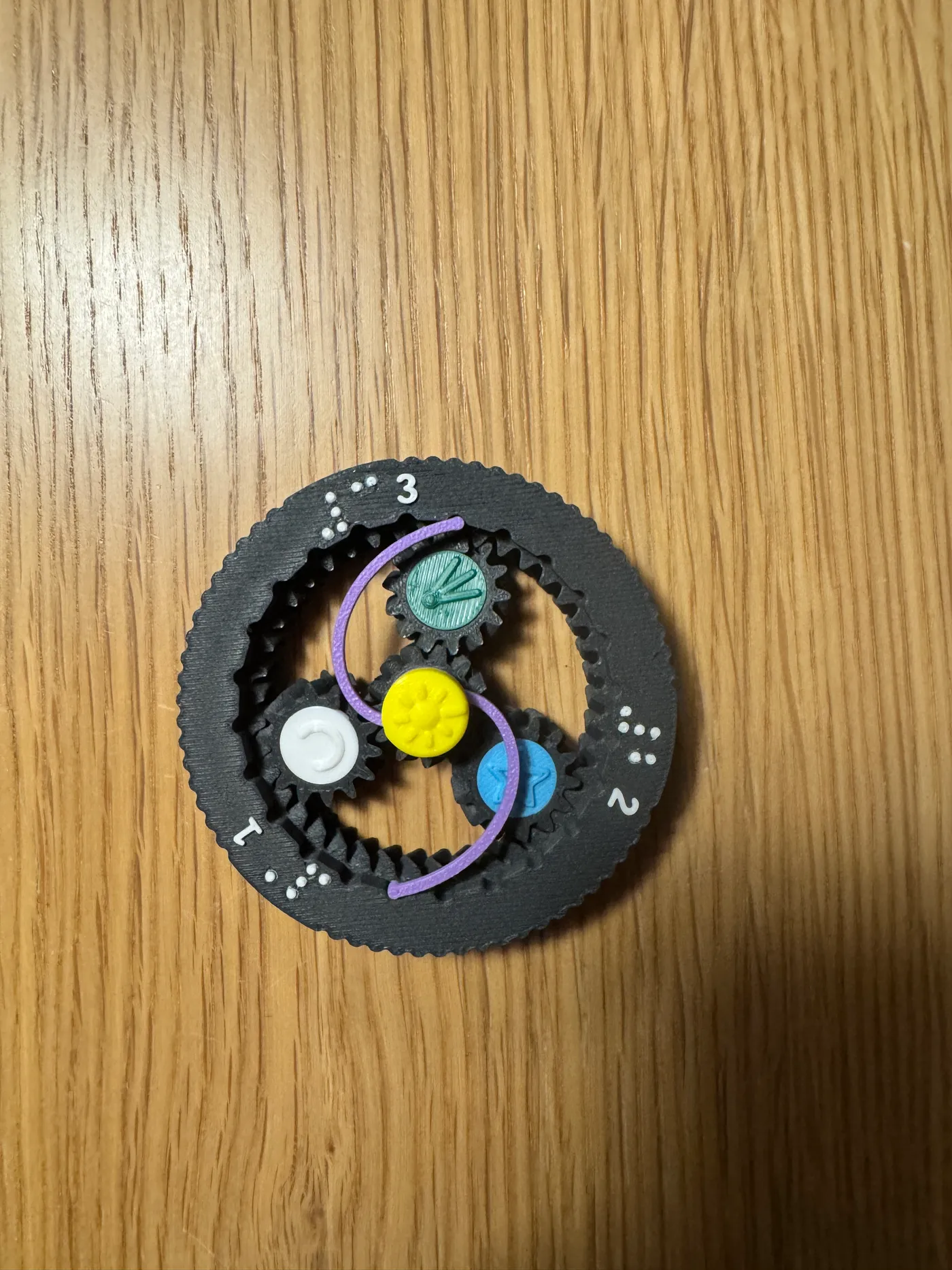 Top view photo of the finished ORBIT spinner on a wooden table: black ring with white raised numerals and braille, yellow sun cap in the centre, white moon, green comet and blue star planet caps, and the purple click spring arms curving between them.