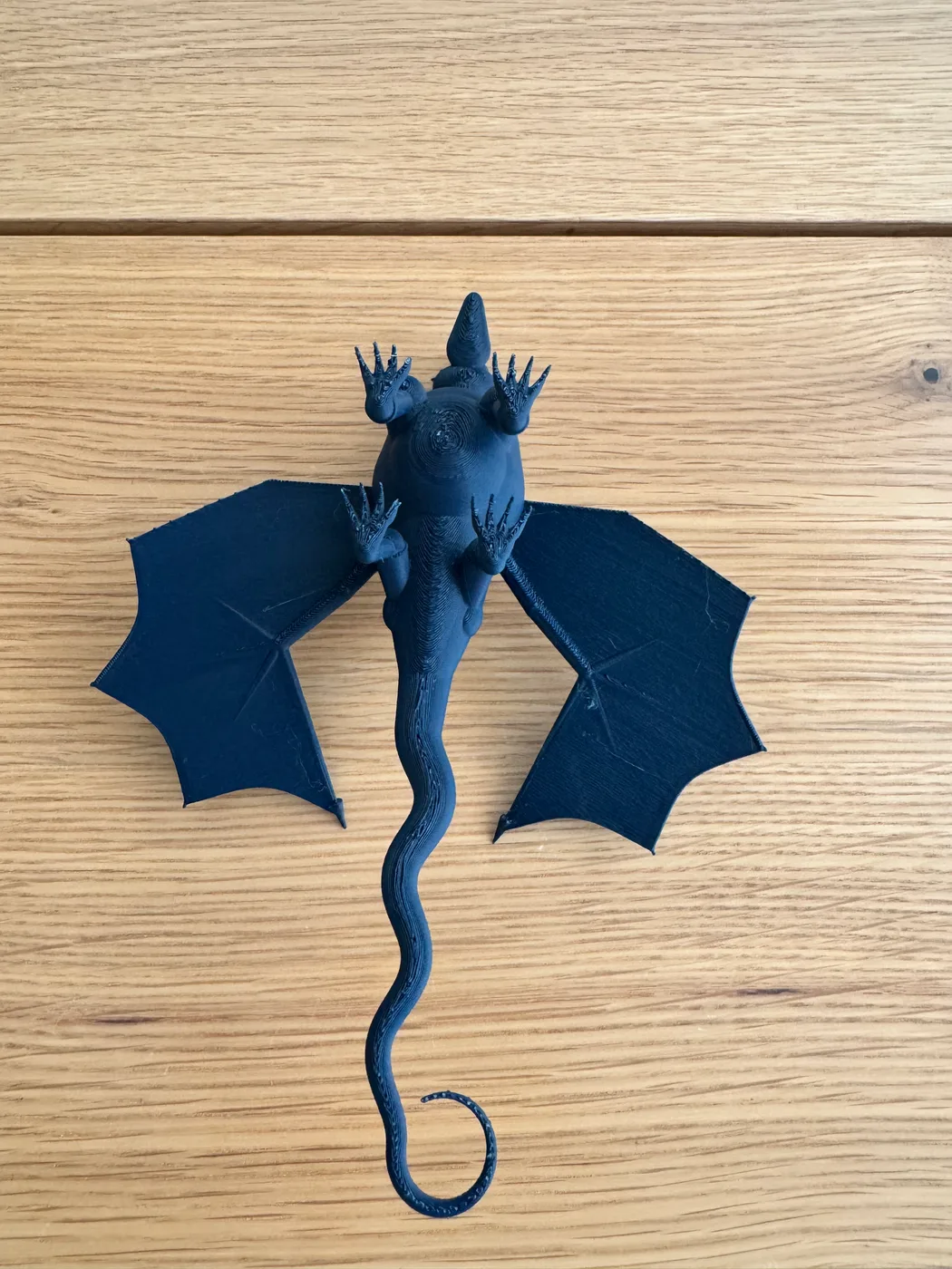 A dark navy 3D-printed dragon lies on a wooden surface, with a horned head, two outstretched bat-like wings and a long tail curling to one side.
