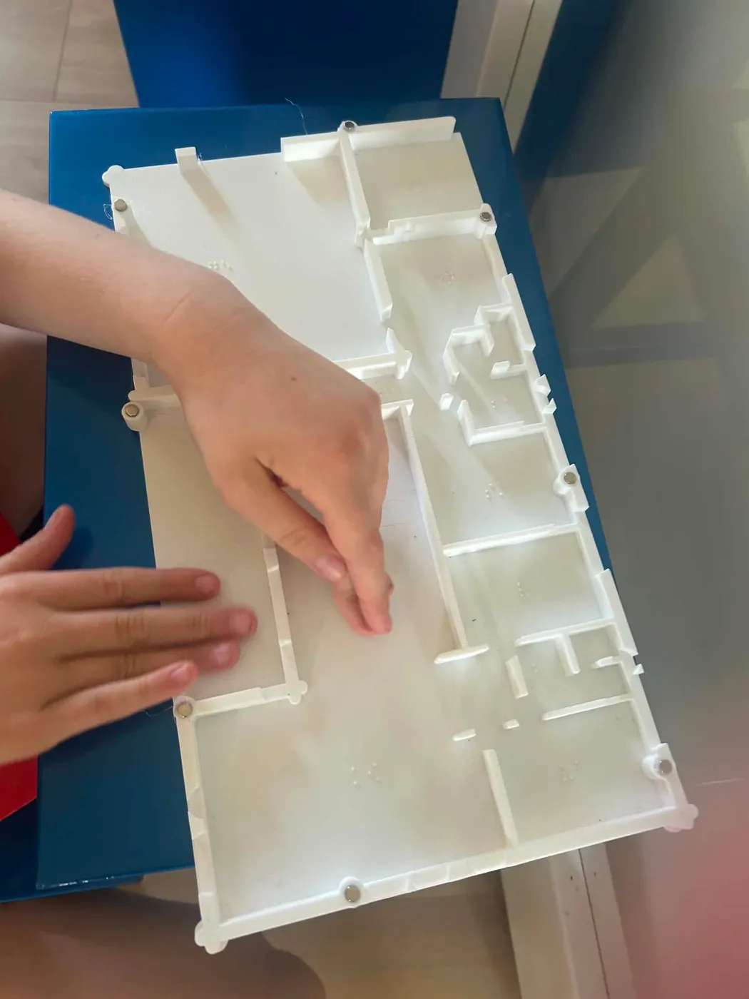 A child's hands reading a white 3D printed tactile floor plan of a home, with raised walls, open doorways and small braille labels in the rooms.