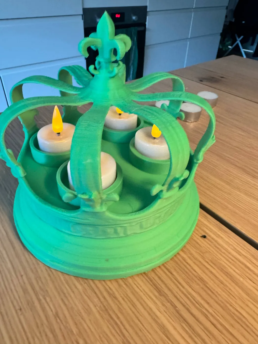 A green 3D printed crown candle holder on a wooden table with four flickering LED tealights glowing between its arches, and spare LED tealights beside it.